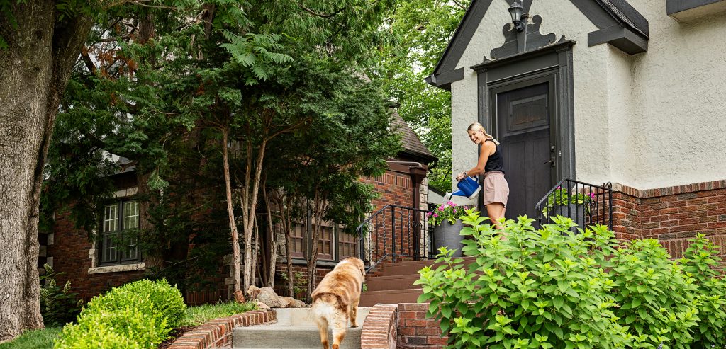 a woman waters flowers on her front porch while a dog walks up the front steps