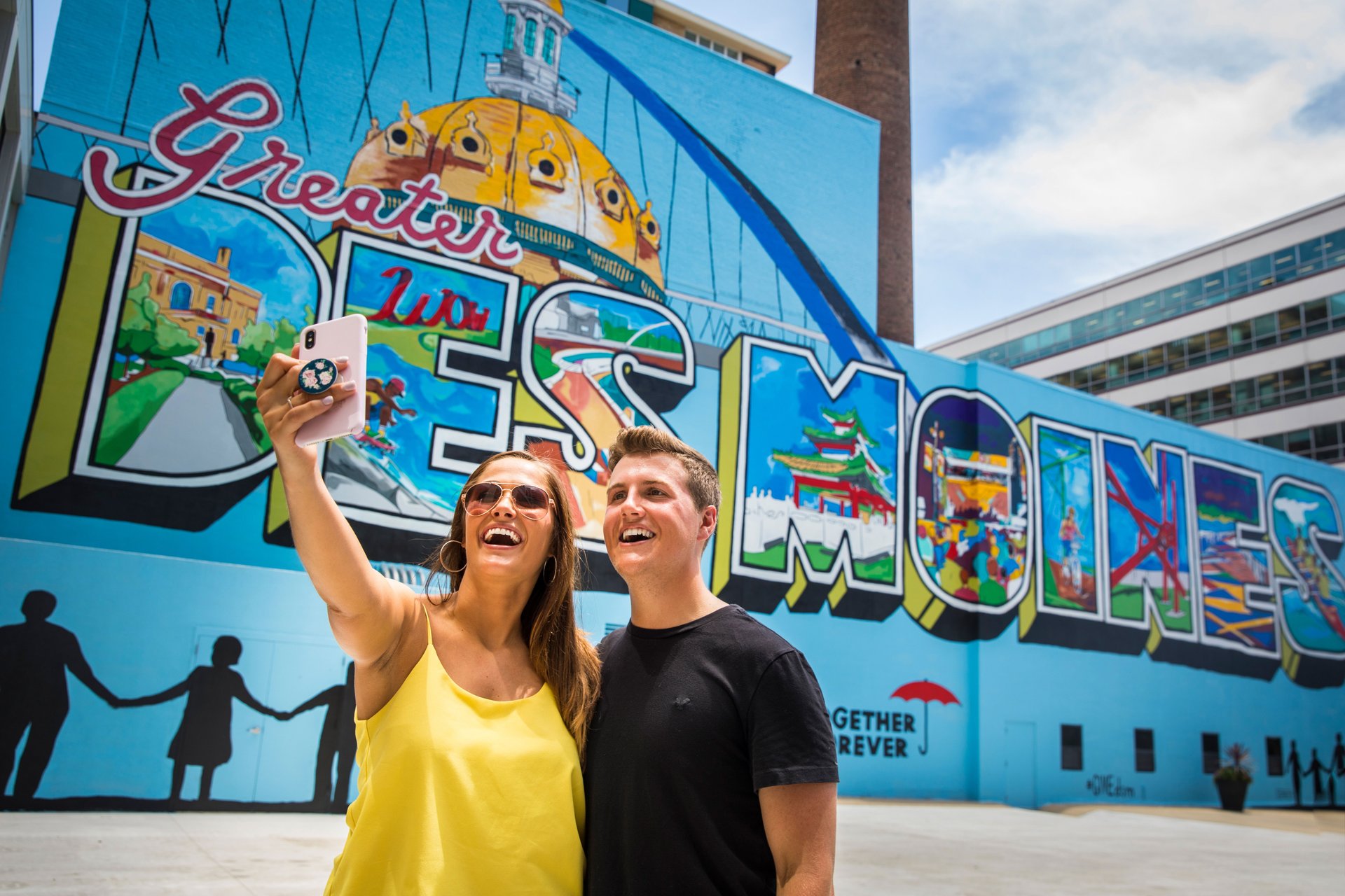 Two people take a selfie in front of a Des Moines mural