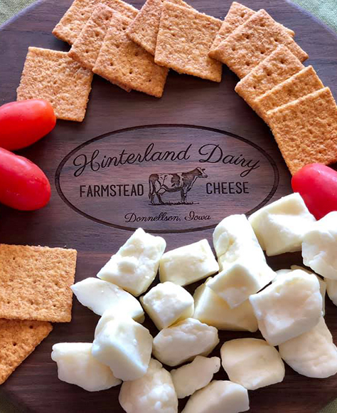 Hinterland Dairy's logo engraved on a wood cutting board surrounded by cheese curds, crackers, and grapes.