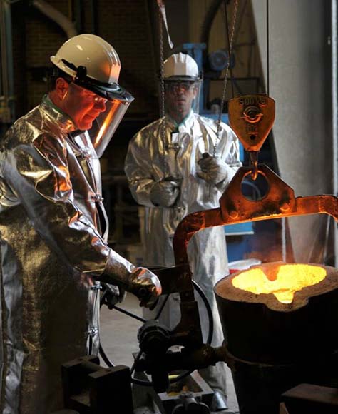 Two men in heat protection gear standing over metal casting.