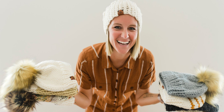 Woman in a knit hat standing with a pile of knit hats on each hand. 