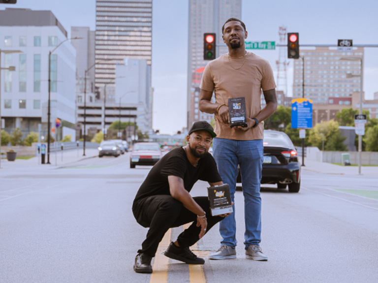 Pernell Cezar Jr. and Road Johnson standing in the road holding boxes of BLK & Bold coffee. 