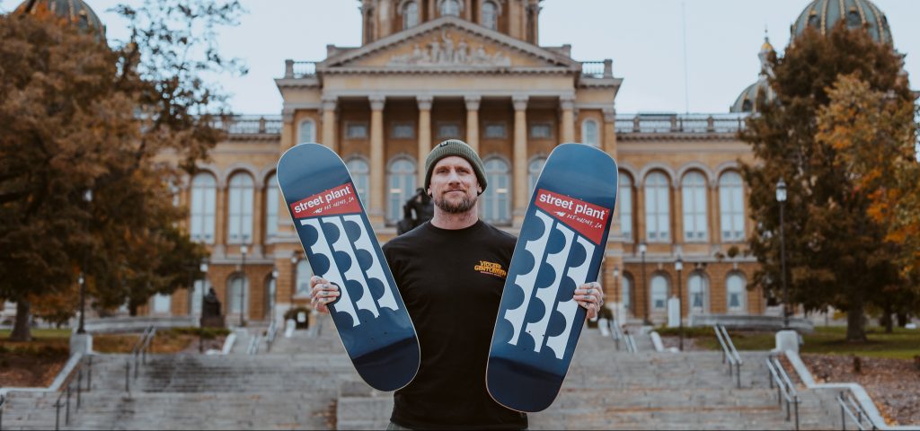 man holds two skateboards in front of Iowa Capitol building