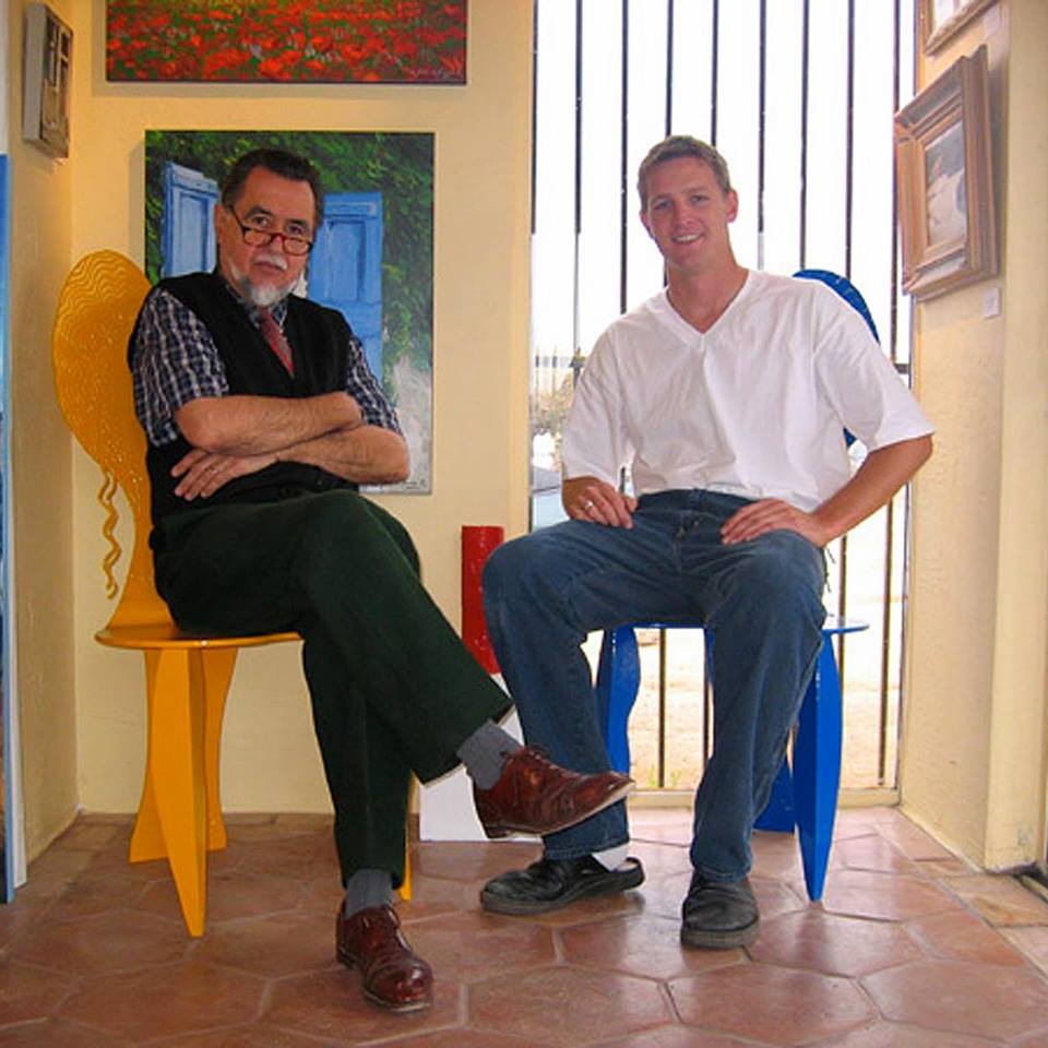 Zach Jones and Sergio Ladron de Guevara sitting in front of a wall lined with original art work. 