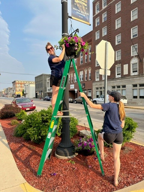 two women using a ladder to hang plants in downtown Marshalltown