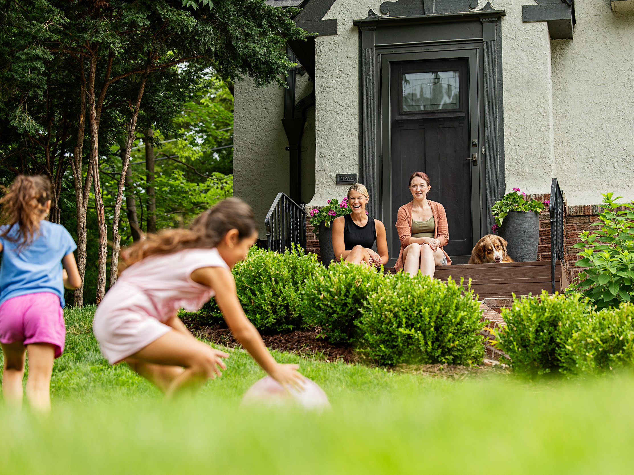 Two moms and a dog sit on the front steps of a house while kids play in the front yard