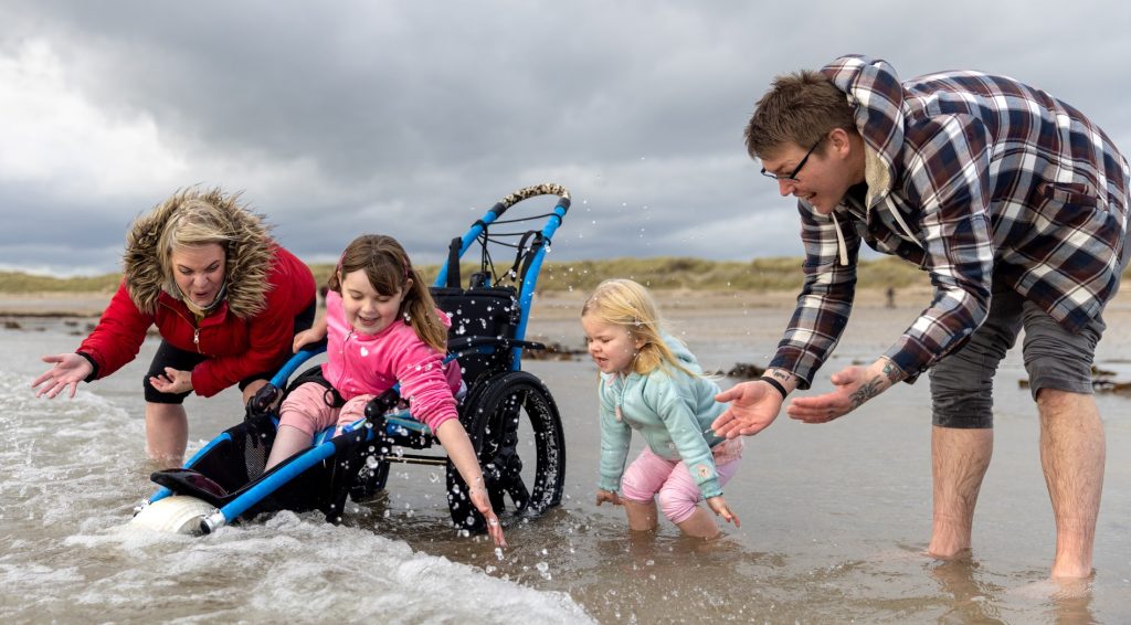 A young girl in a wheelchair and her family splash in the water at a beach.