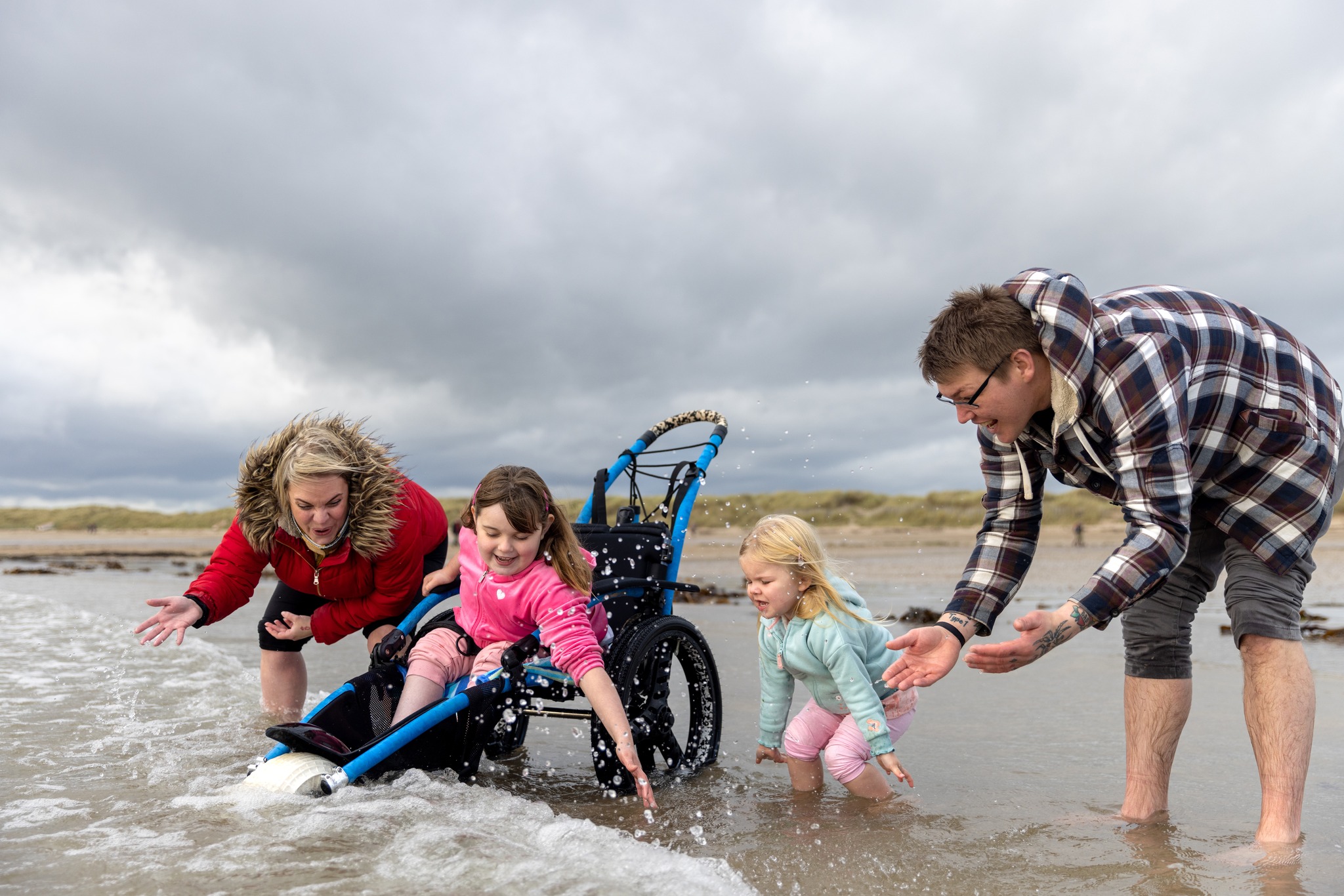 A child in a wheelchair and her family splash in the water at a beach.