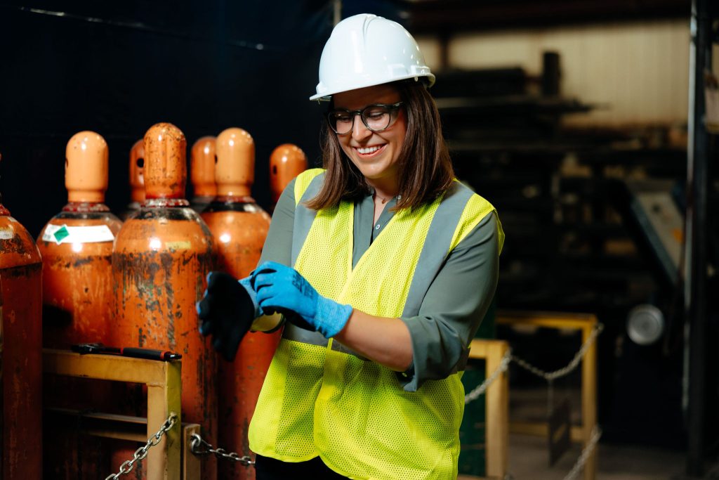 A woman wearing a safety vest and helmet smiles while putting on work gloves.