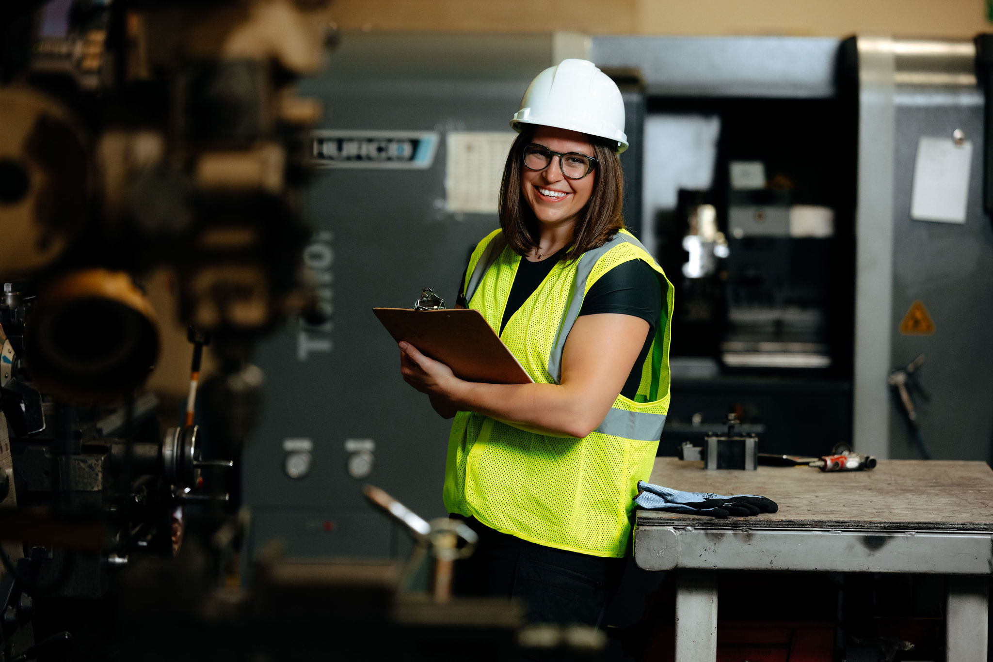 A woman wearing a safety vest and helmet smiles while holding a clipboard.