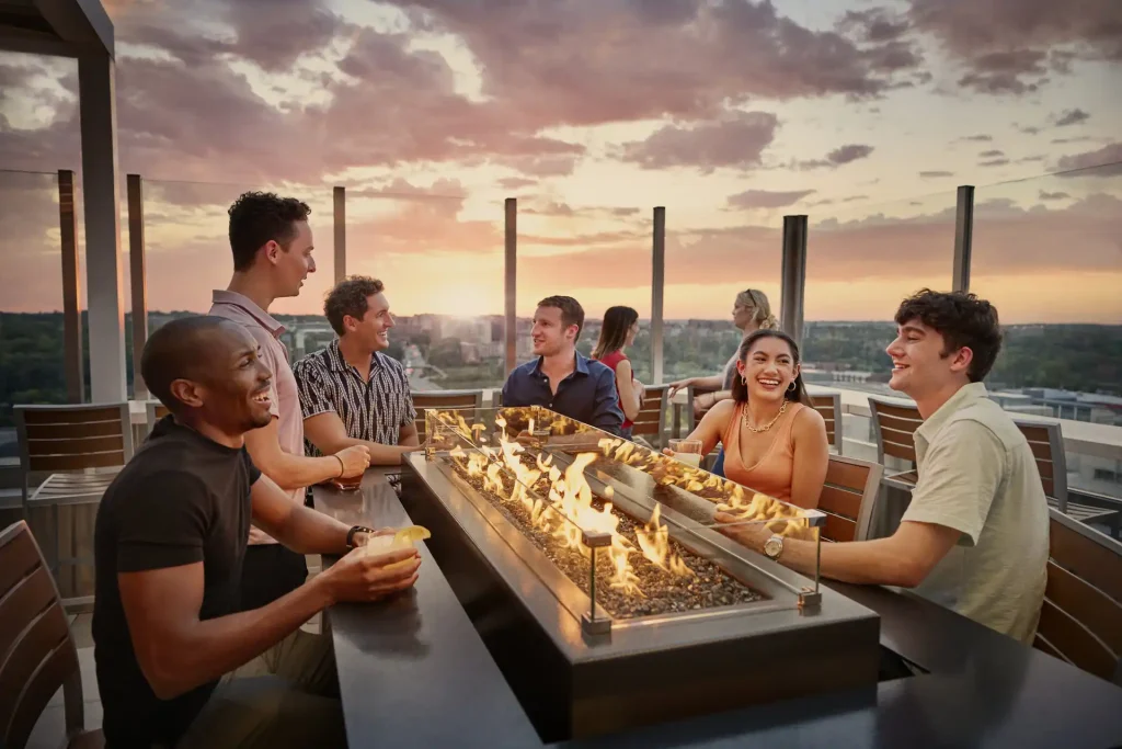 group of people sipping drinks around an outdoor tabletop fire, against a city skyline