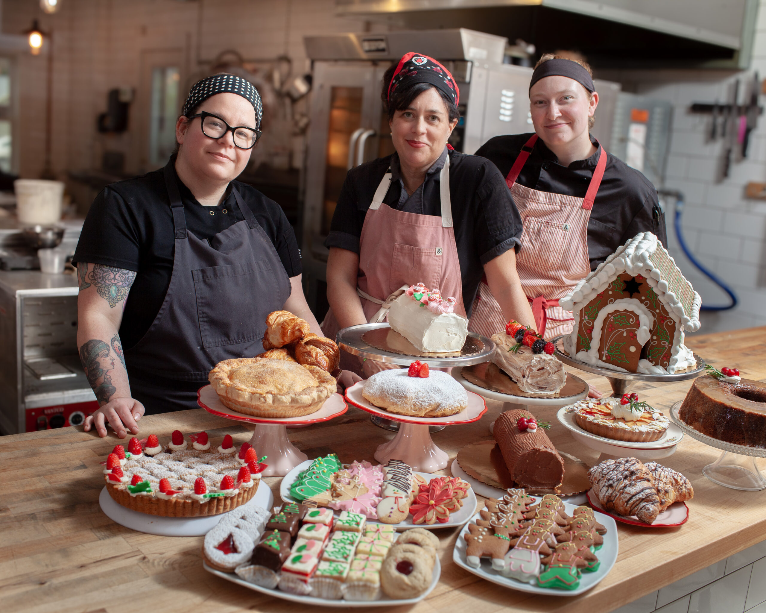 Jamie Powers and two coworkers stand behind a wooden table covered with various decorated cakes, cookies, pies, and pastries in a bakery kitchen.